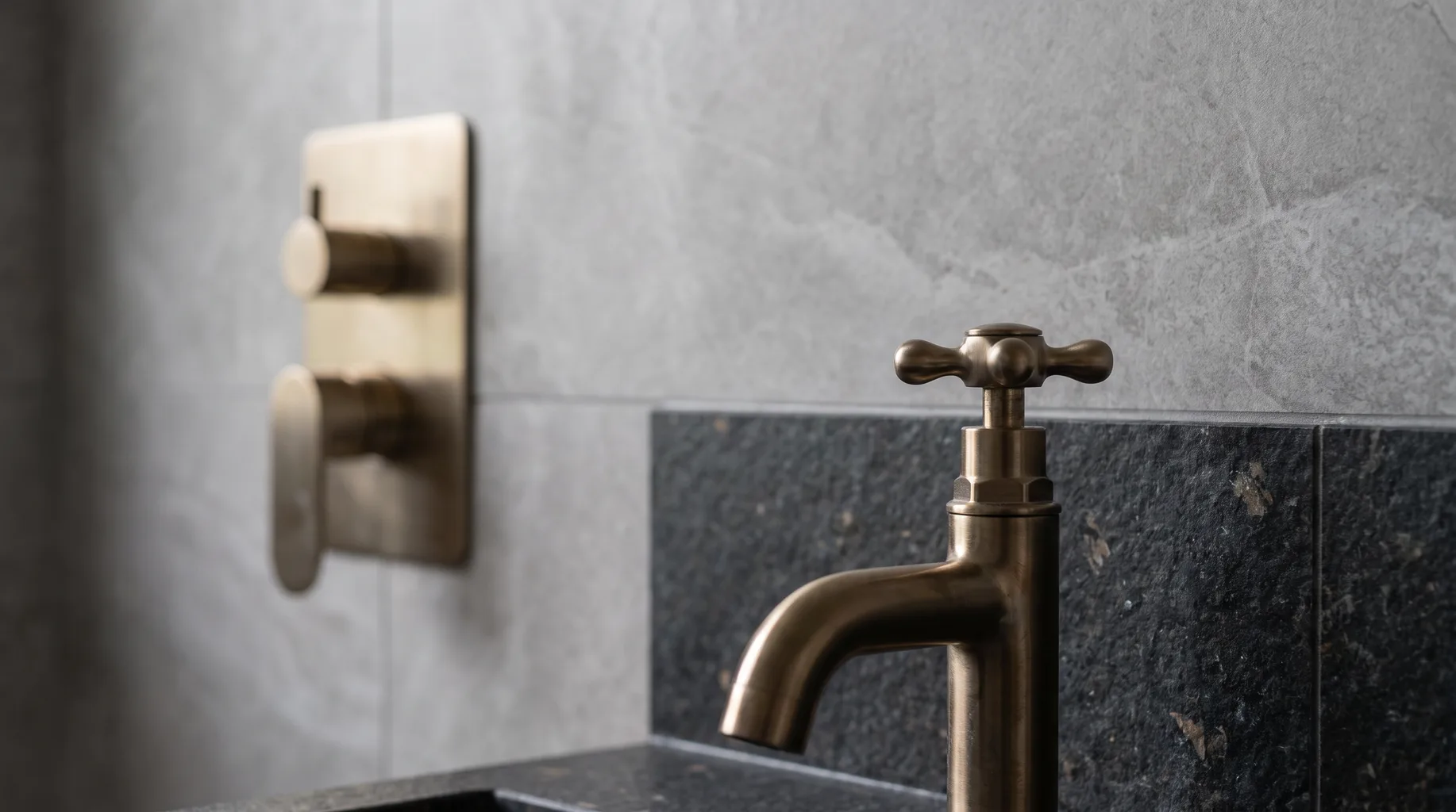 Close-up of a traditional brushed bronze cross-head basin tap beside a contemporary concealed shower control panel on dark basalt stone tiling in a Wokingham bathroom