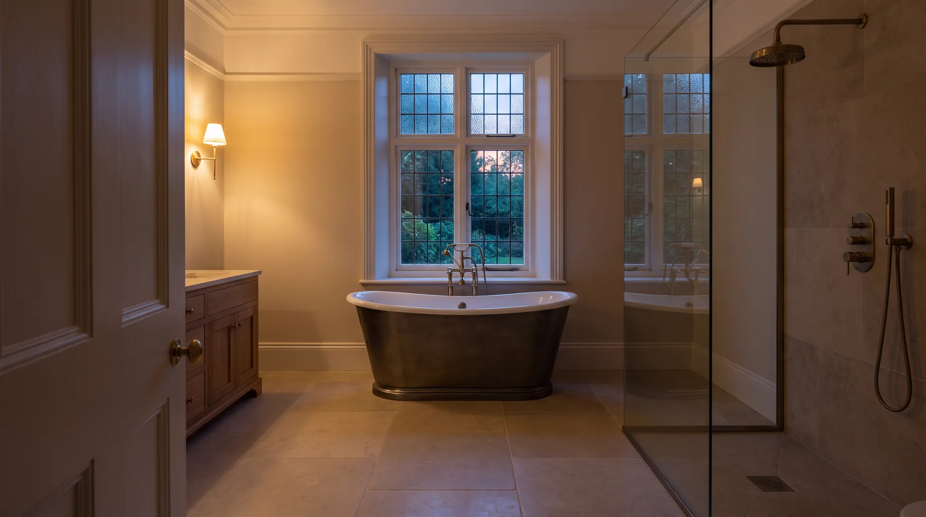 A finished Ascot bathroom with a freestanding bath beneath a tall window, honed limestone floor and brushed bronze fittings, walk-in shower visible beyond a glass partition in warm evening light.