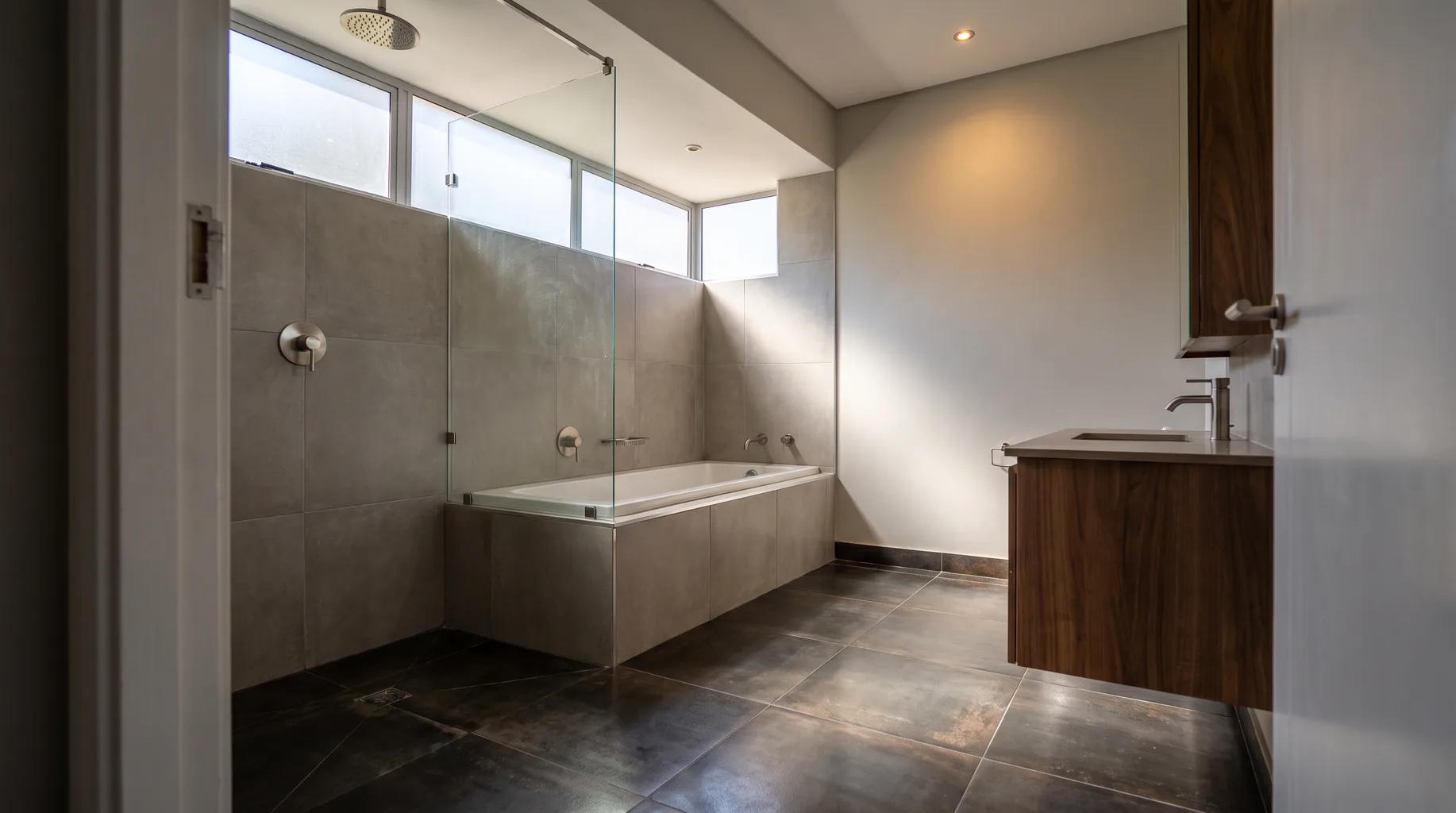 A finished Sunninghill family bathroom with a tiled shower bath, dark basalt-effect floor tiles, brushed nickel fittings and a walnut vanity unit under bright filtered clerestory daylight.