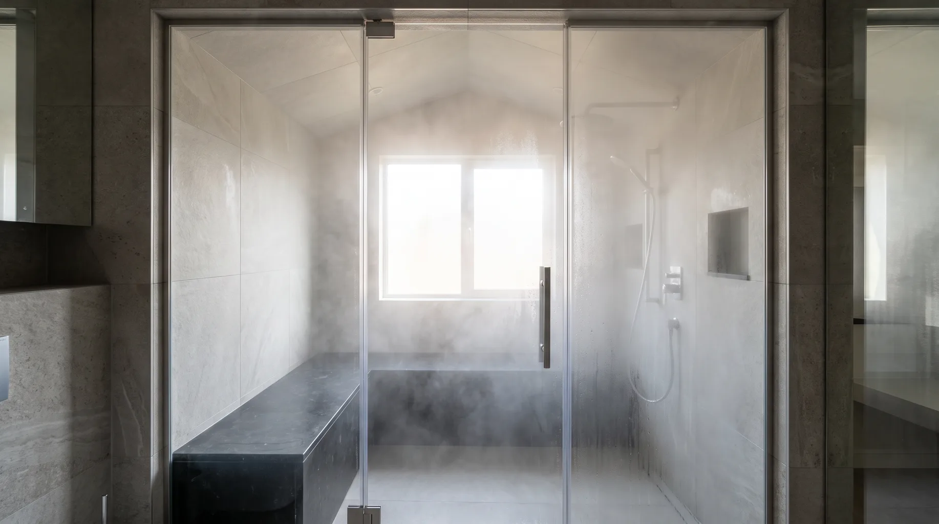 Finished steam shower enclosure with a sealed full-height glass door, tiled bench and sloped ceiling, filled with steam haze