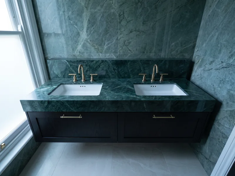 Finished double vanity unit with two inset basins and brushed brass taps against a deep green marble wall in a wide-angle bathroom view
