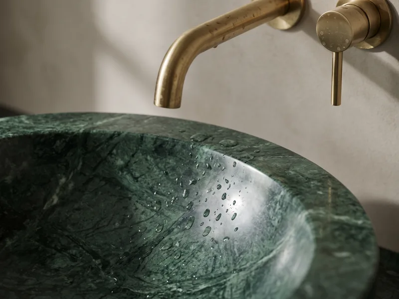 Close detail of a polished deep green marble countertop basin with a brushed brass tap, water droplets beaded on the stone surface