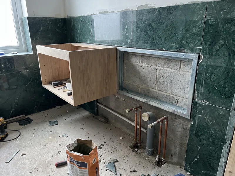 Part-built wall-hung vanity carcass fixed to a concealed steel frame against a deep green marble-tiled bathroom wall, first-fix pipework visible below