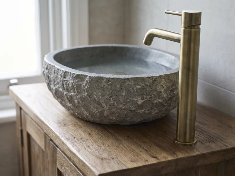 Close detail of a hand-carved stone vessel basin with a brushed brass pillar tap on a timber-topped vanity