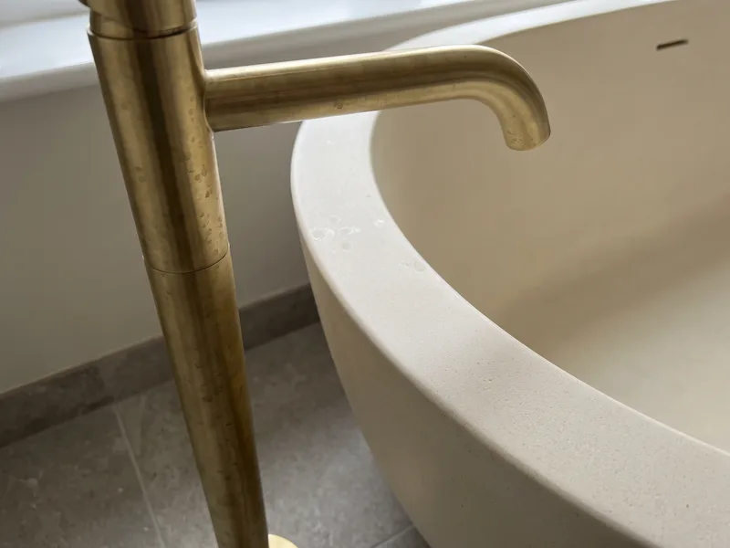 Close detail of a tall brushed brass floor-standing bath filler tap beside the rolled edge of a stone-resin freestanding bath