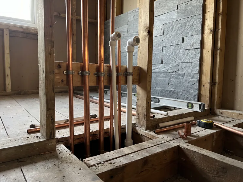 Close detail of copper and plastic first-fix pipework rising from a subfloor against a part-built travertine stone bathroom wall
