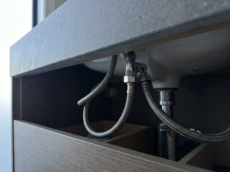 Close detail of brushed bronze flexible hose connections beneath a travertine stone vanity countertop basin
