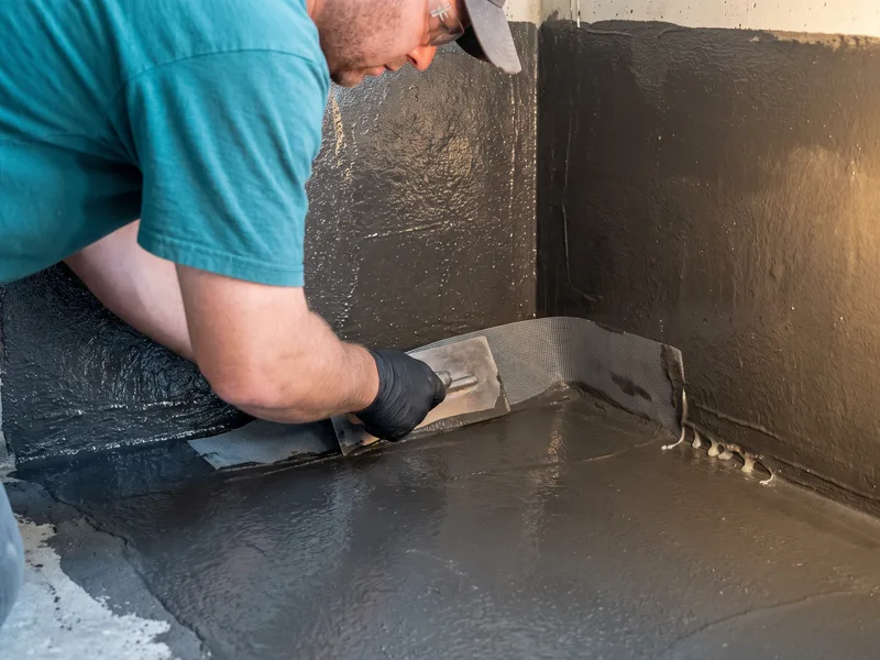 Installer bedding waterproof reinforcement tape into a liquid tanking membrane at a floor-to-wall corner junction