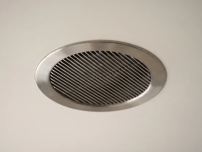Macro detail of a circular brushed-metal extractor fan grille set flush and finished seamlessly into a painted ceiling