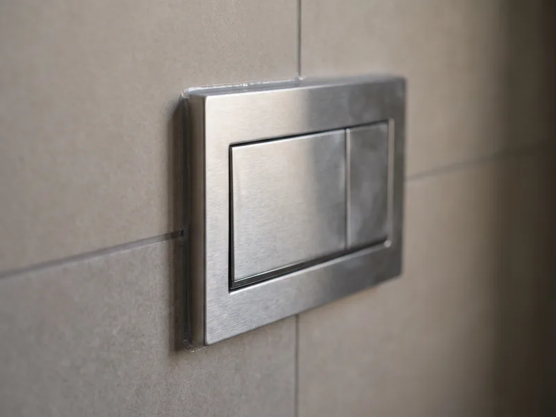 Macro detail of a brushed-metal dual-flush plate set flush into fully grouted large-format porcelain tiling