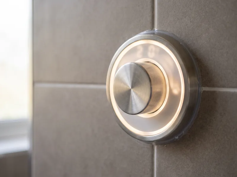 Macro detail of a brushed-metal digital shower control pad with illuminated display ring, fixed flush to large-format porcelain tiling