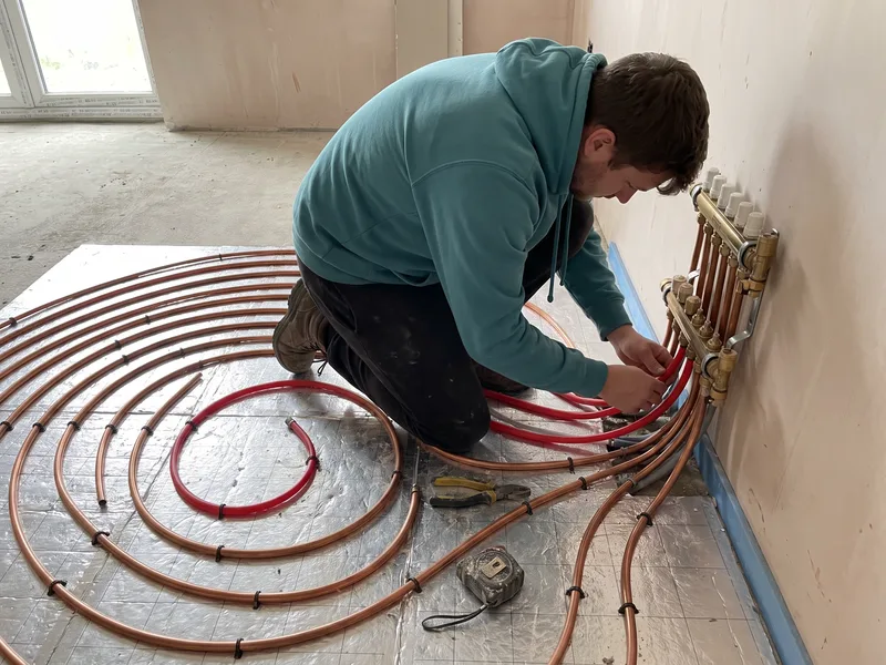 Installer connecting a wet underfloor heating pipe loop to a wall-mounted manifold before screed is poured
