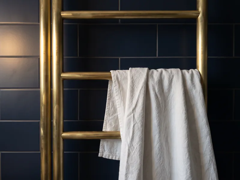 Close detail of a brushed brass vertical heated towel rail on a navy tiled wall with a white towel draped over it