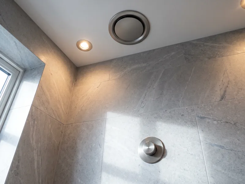 Close detail of a flush ceiling extract grille beside a recessed downlight, with a satin pewter humidistat control on a honed dove-grey limestone wall below