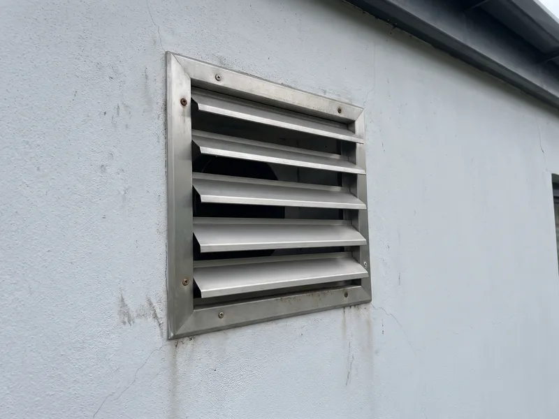 Close three-quarter detail of a satin pewter louvre extract terminal set into a pale grey rendered external wall