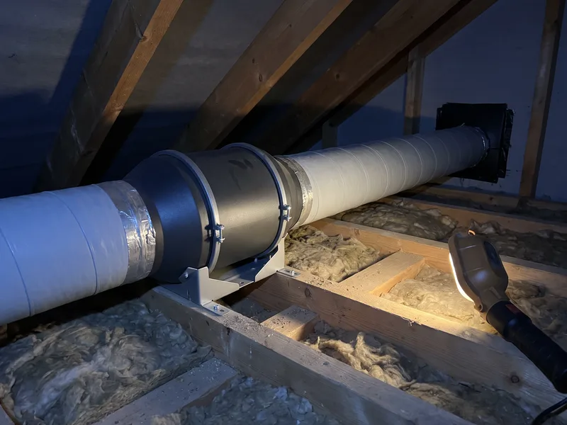 First-fix insulated rigid ducting connected to an in-line centrifugal extractor fan mounted among loft joists, cool grey tones under a single work light