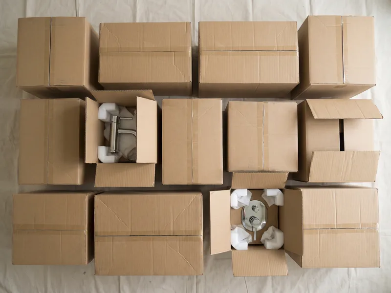 Overhead flatlay of unmarked cardboard boxes of sanitaryware and brassware, sourced directly by the client under a fit-only arrangement