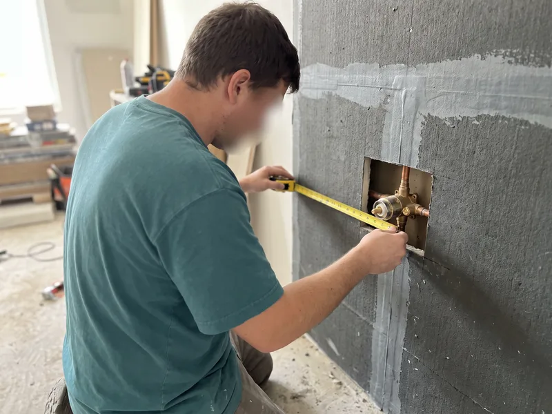 Installer measuring the depth of a recessed shower valve body against a backer-board wall before tiling