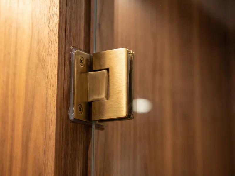 Close detail of a brushed brass hinge and bracket fixing a glass shower door to a warm walnut-panelled wall