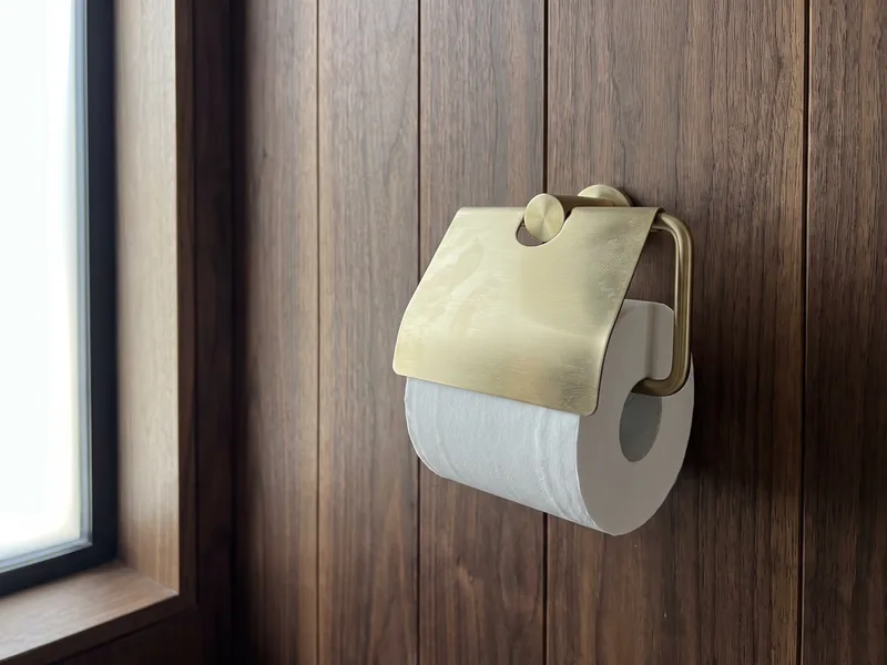 Close detail of a brushed brass wall-mounted toilet roll holder fixed to a walnut timber-panelled wall
