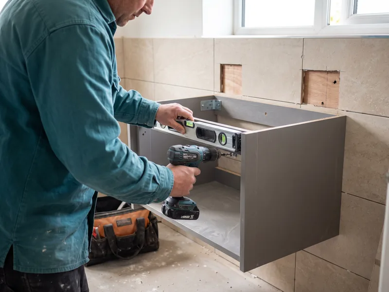 Installer fixing a wall-hung vanity unit carcass to a reinforced wall backing, checking level before the basin is fitted