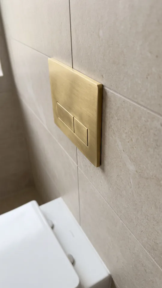 Close-up of a brushed brass dual-flush plate set flush into tile on the duct face above a wall-hung toilet