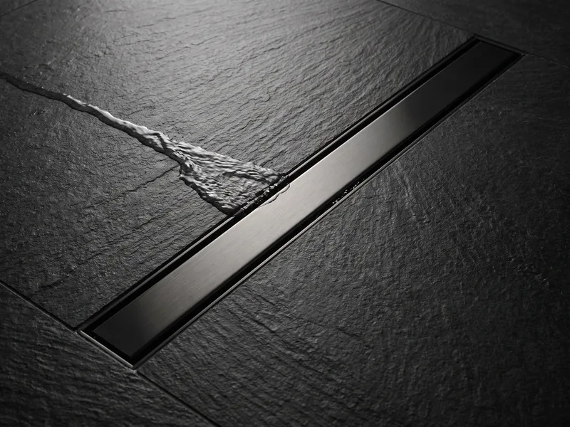 Close detail of a brushed black-nickel linear drain channel set flush into black stone tile with water flowing into it