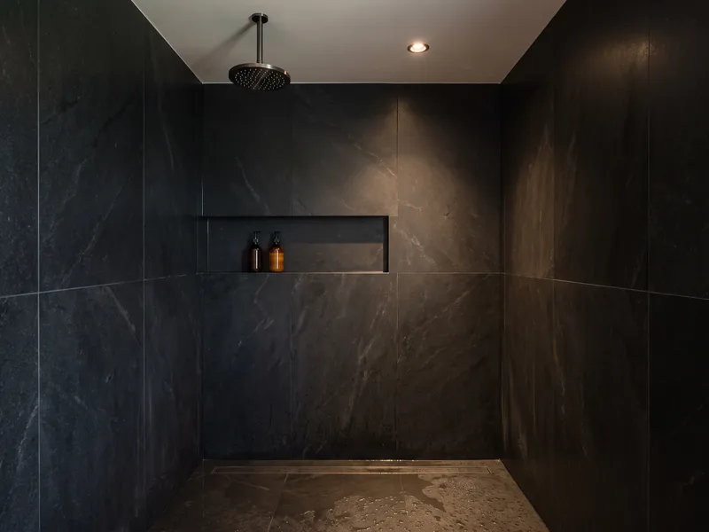 Finished wet room shower zone with an overhead shower arm and recessed niche shelf against black stone walls
